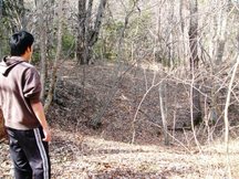 若い男性が茶色いパーカーと黒いジャージ姿で落ち葉の積もった冬枯れの林に立ち、葉の落ちた細い木々の間から奥の斜面の砂金流し掘り跡をじっと見つめている写真