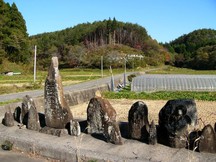 田畑の広がる農村風景の前に苔むした石碑や石仏が並び、背後には山林が見える気仙道の写真