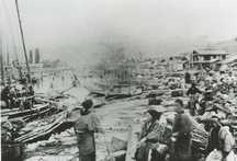 川沿いの港に複数の船が停泊しており、岸には木材や荷物が大量に積まれていて、物流や作業の拠点として活用されている様子が写された古い写真