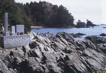 切り立った岩場と穏やかな海を背景に、石垣の上に建てられた複数の石碑が写っている写真