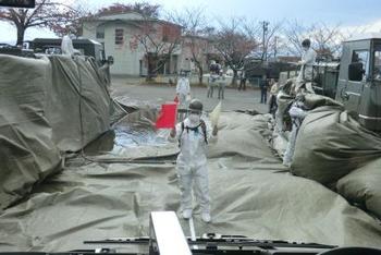 白い防護服とマスクを着用した人物が立ち、両手にそれぞれ赤と白の旗を持って避難車両を誘導している写真