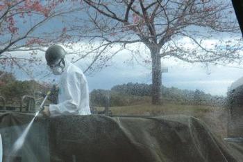 防護服とヘルメット、マスクを着けた人物が、高圧洗浄機のような機械で車両かシートの表面を洗浄している写真