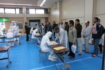 広い室内には青いビニールシートが床一面に敷かれ、白い防護服を着た人たちの前に作業服や私服の人々が列を作り並んでいる写真