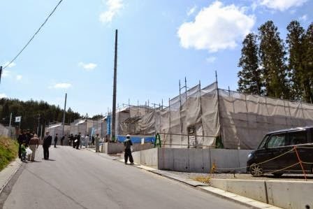 晴れた空の下、餅まきが終わり人が居なくなった緩やかな傾斜のある道路沿いに建てられた建設中の複数の住宅が、足場に囲まれ整然と並んでいる写真