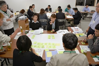 黄色の付箋がたくさん貼られた大きな模造紙が置かれた机を囲んで、男女5人が座り、周りに3人の男性が立って模造紙を見ている話し合いの様子の写真