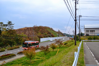 道路沿いにバスが走っており、背後に盛土と白いフェンスが続きいている造成工事道路沿いの写真