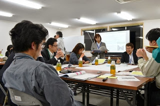 中央に模造紙が置かれた机の上に資料を広げ、男女のグループが話し合いをしている様子の写真