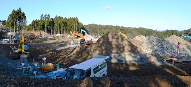 業務用のバンや建設資材が並び、多数のショベルカーが岩や土を動かす広い造成工事現場の写真