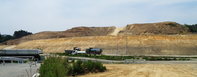 整地された斜面の下に大型トラックが並び、造成が進んで山の形がなだらかになった工事の途中段階の様子の写真