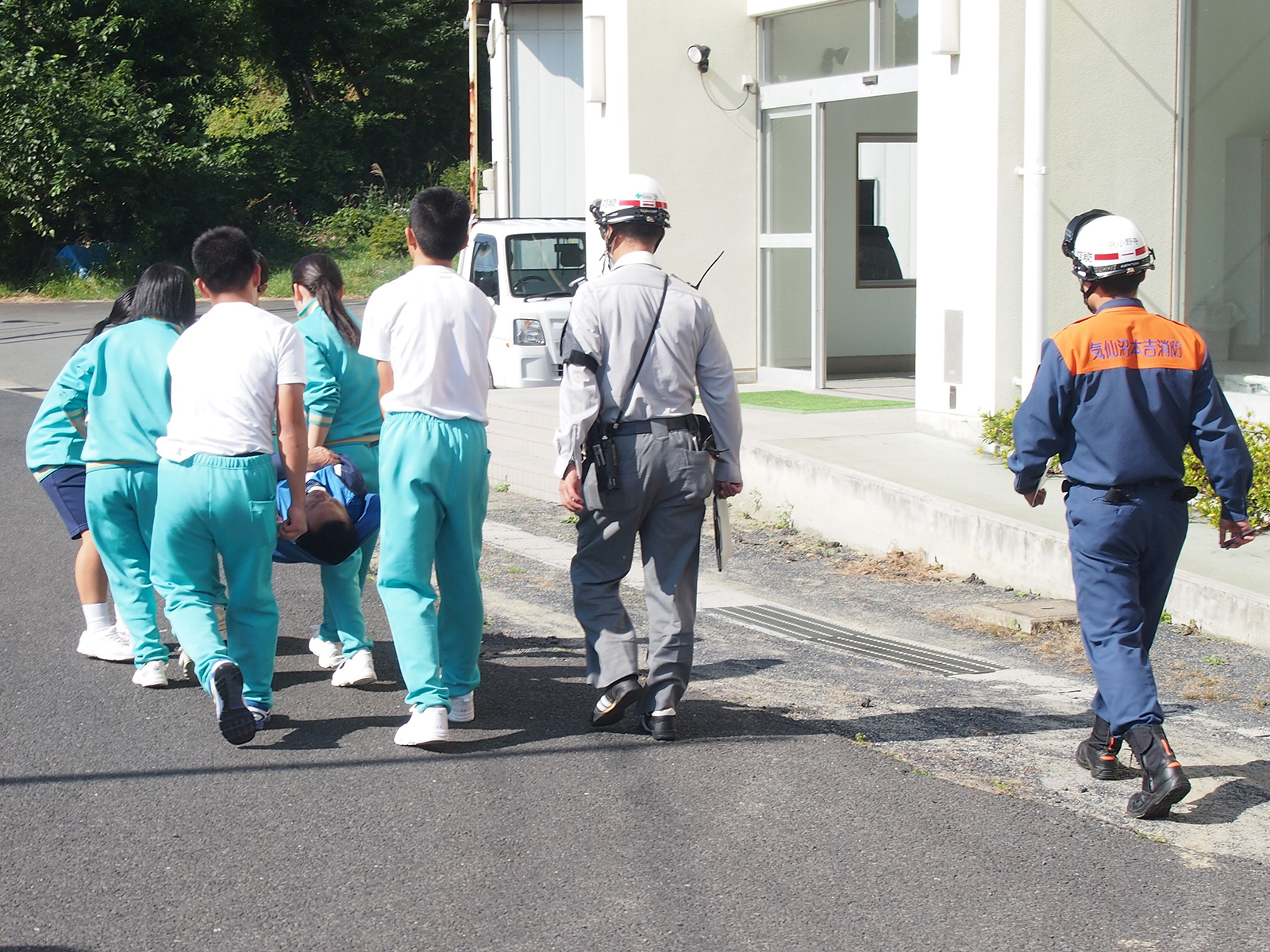 ジャージ姿の高校生5人が簡易的な担架でけが人役の男性を運び、消防隊員らと共に避難訓練を行っている後ろ姿の写真