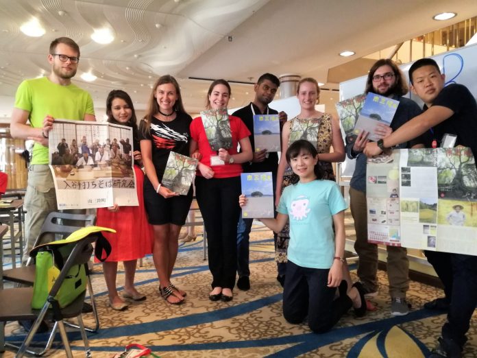 多国籍の若者たちが室内で笑顔で立ち並び、手にパンフレットやポスターを持ちながら、異文化交流イベントの成果を紹介している様子の写真