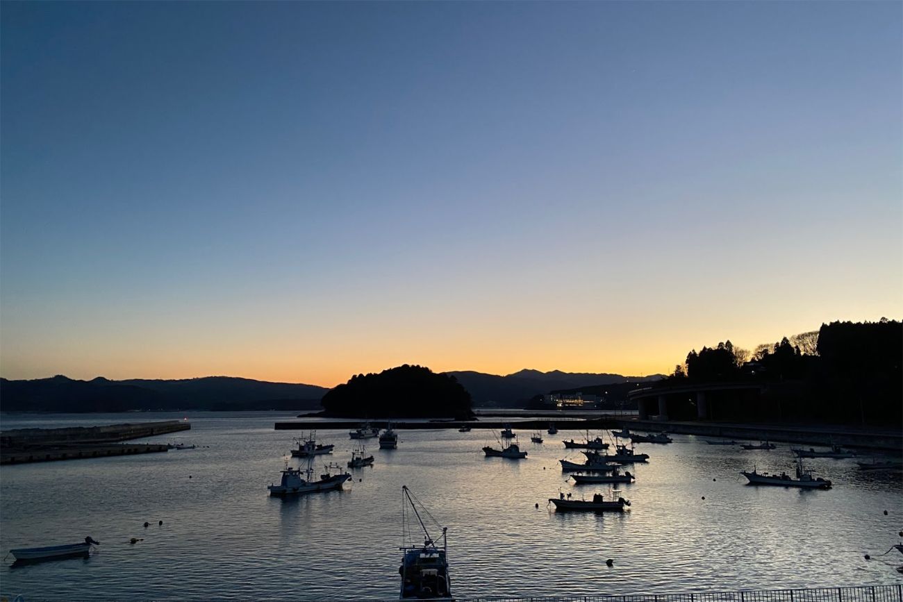 日が沈みかけた薄明かりの空の下、静かな湾に小さな漁船が点在し、遠くの島や山々のシルエットが映る穏やかな港の写真