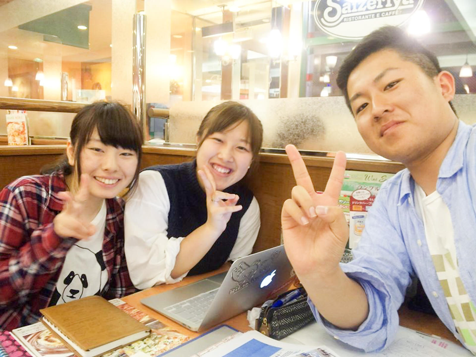 ファミリーレストランのテーブル席でノートパソコンや資料を広げテーブルを囲んだ佐々木さん、佐藤さん、西城さんが笑顔でピースサインをしている写真