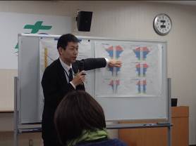 ホワイトボードに貼った6つのグラフが載った紙を指しながら、マイクを持って話をしている男性の写真