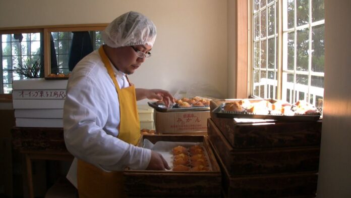 不織布キャップを被り白衣と黄色いエプロンを着たパティシエの男性が、窓側に積まれた木箱に並んだマドレーヌを手作業で整えている工房内の写真