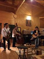 木の壁に囲まれた空間で、3人の男性がギター、ウッドベース、サックスを演奏している写真