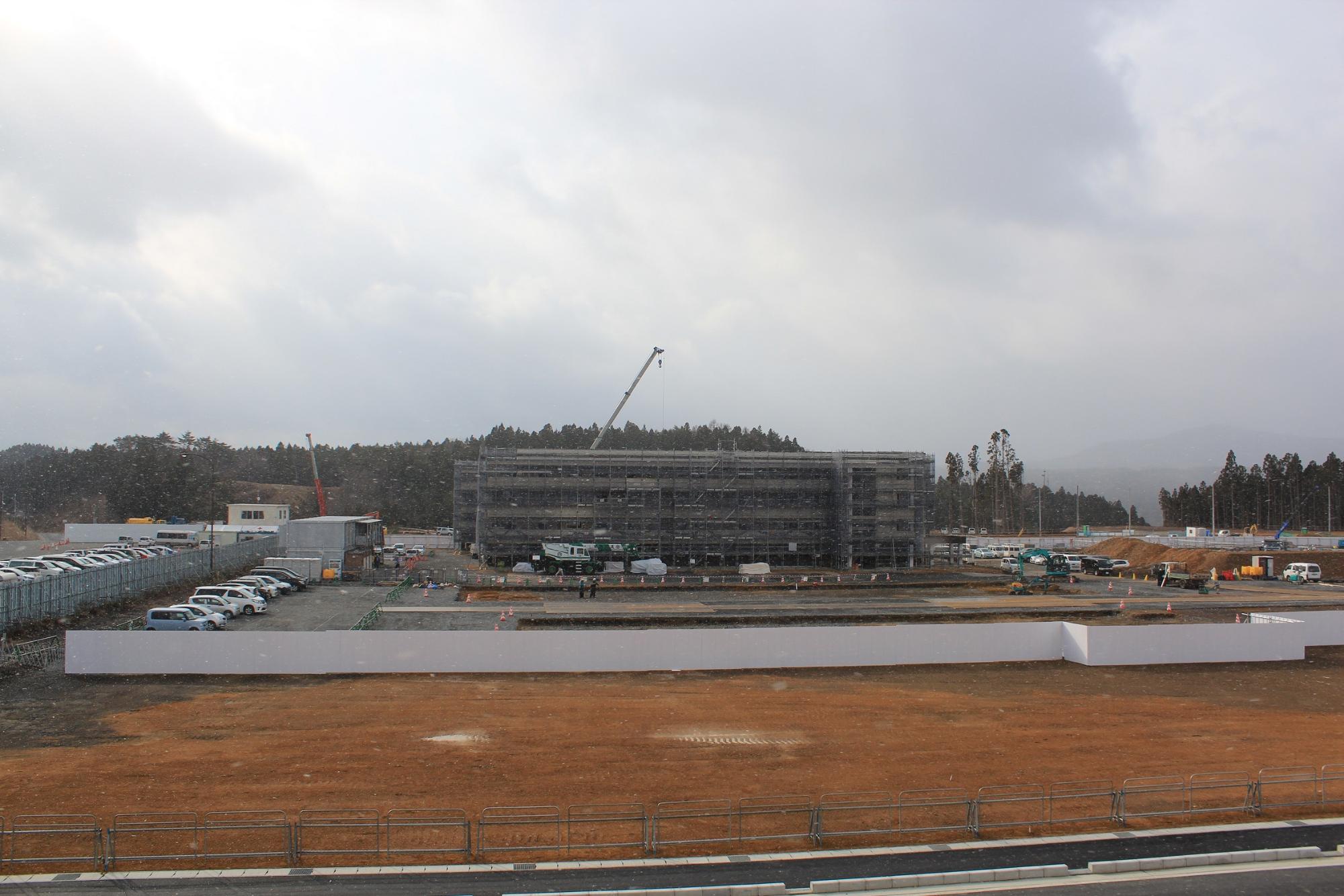 曇り空の下、うっすらと雪が舞うなか、中央に足場に覆われた建設中の建物とクレーンがあり、その手前には整備された駐車場や作業エリアが広がっている写真