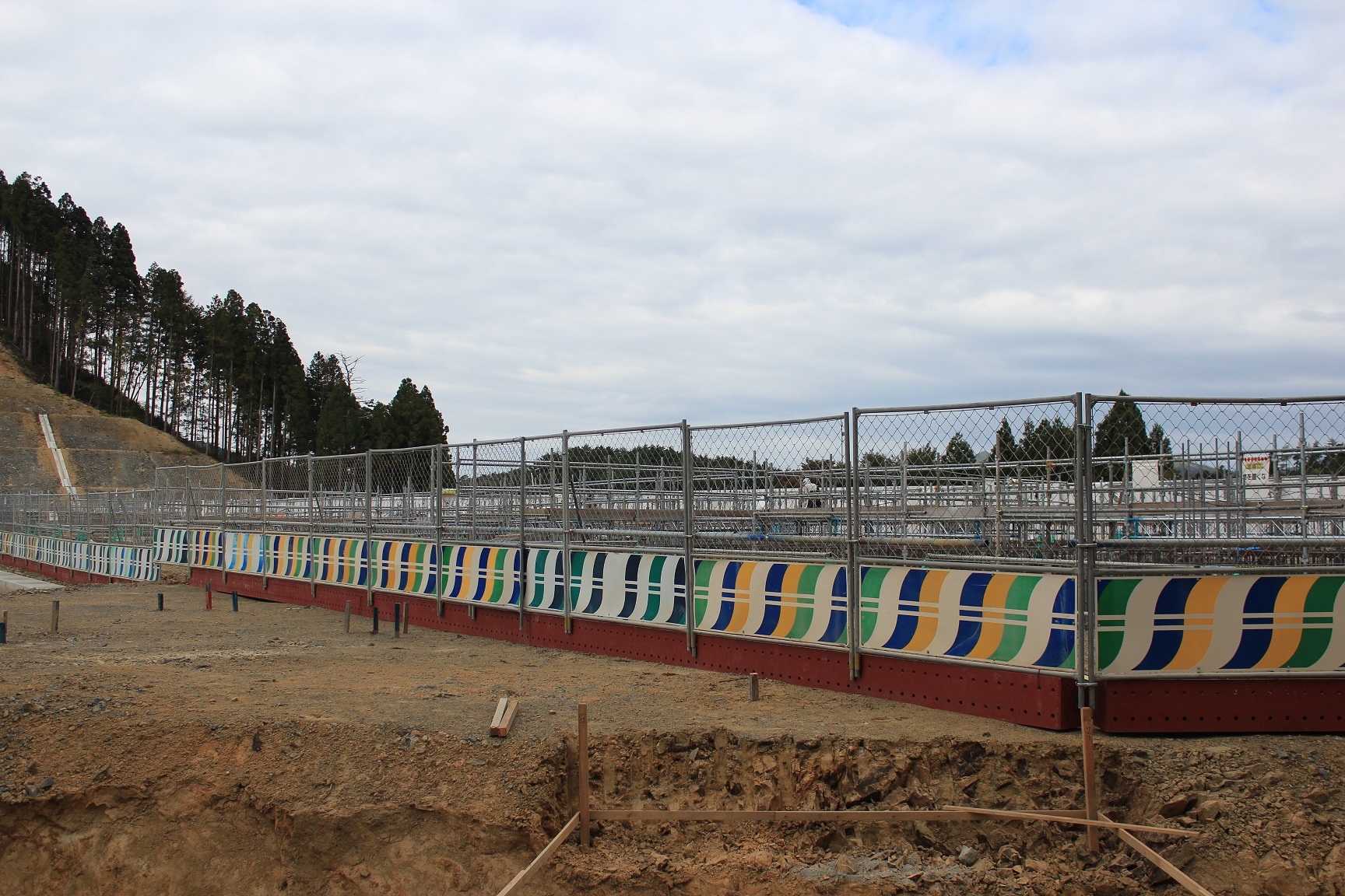 曇り空の下、カラフルな模様が描かれた安全フェンスに囲まれた造成地では、基礎工事が進行中で、背景には整備中の斜面と林が広がっている写真