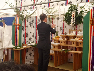 供物や榊が並んだ神聖な祭壇の前で、黒いスーツ姿の男性が手を合わせて拝礼している後ろ姿の写真