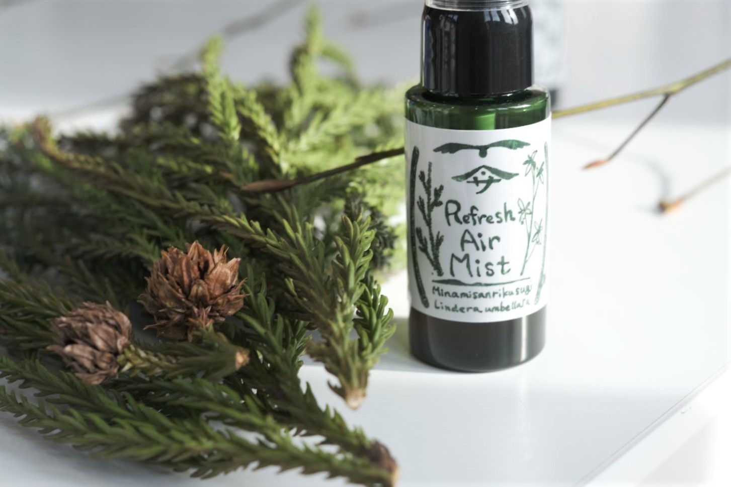 クロモジの枝と杉の葉と「Refresh Air Mist（リフレッシュエアミスト）」と書かれたラベルの緑色のスプレーボトルの写真