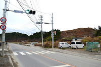 信号機や歩道のある右側に工事事務所や車が並び、背後に木々が伐採された造成前の山が見える工事初期の様子の写真