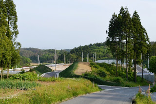 緑に囲まれた道の先に土が盛られた区画と新しい舗装道路が見える郊外の風景の写真