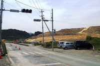 道路脇に車とカラーコーンが並び、山がなだらかに削られている造成工事が進行中の様子を信号機のある道路側から撮影した写真