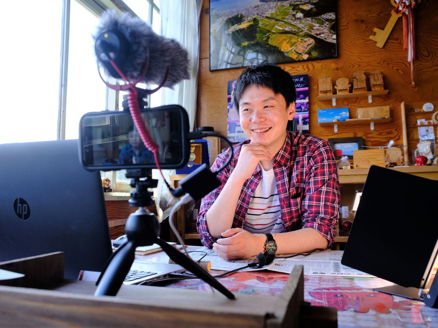 木の工芸品や写真が飾られた部屋で、チェックシャツを着た大森さんが机に向かって笑顔でオンライン配信をしている様子の写真