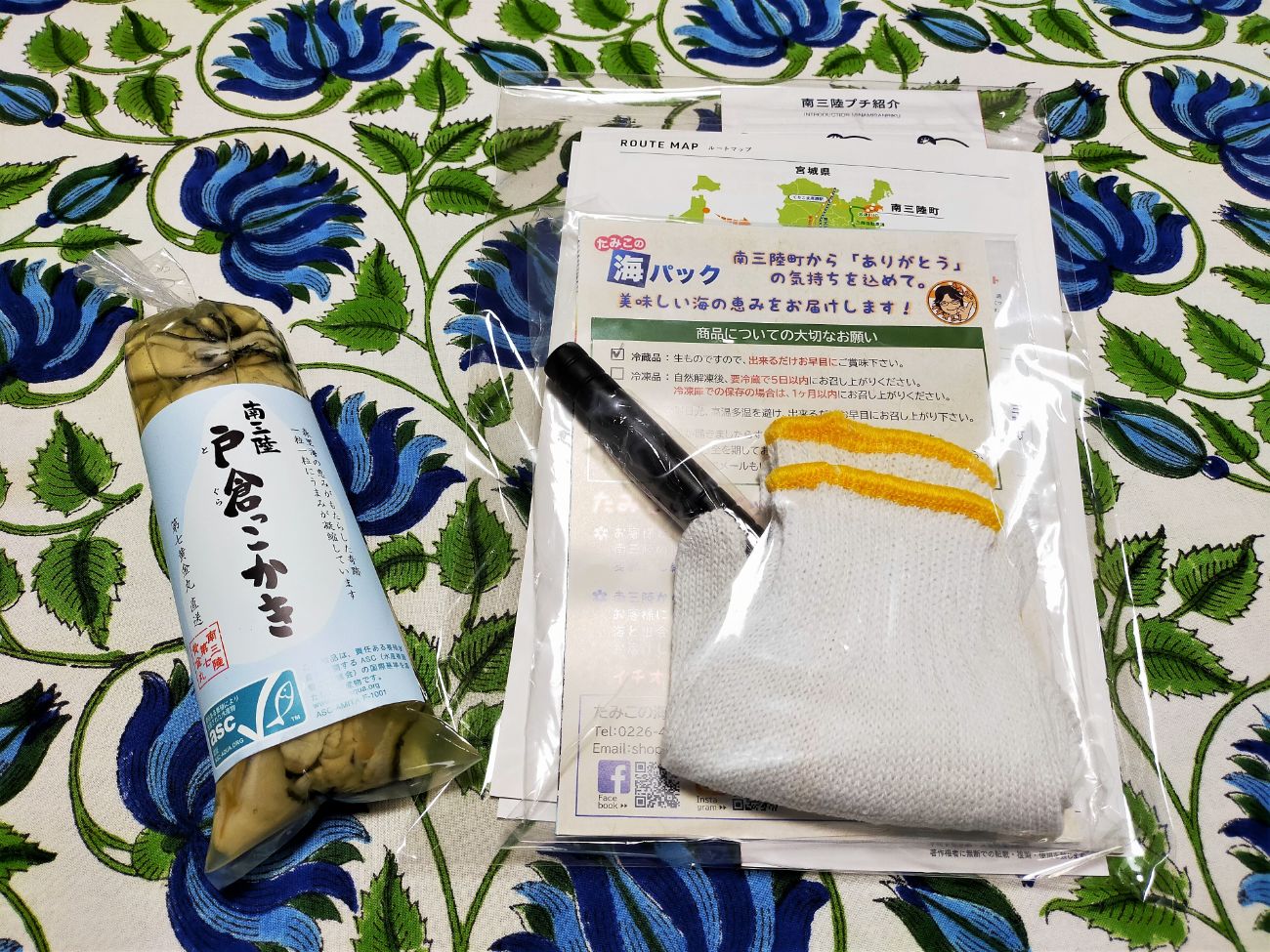 青い花柄のテーブルクロスに、左側に中身が見える透明袋のパッケージに入った牡蠣の加工品が置かれ、右側には軍手と牡蠣ナイフ、案内用のパンフレットが透明の袋にまとめられている写真