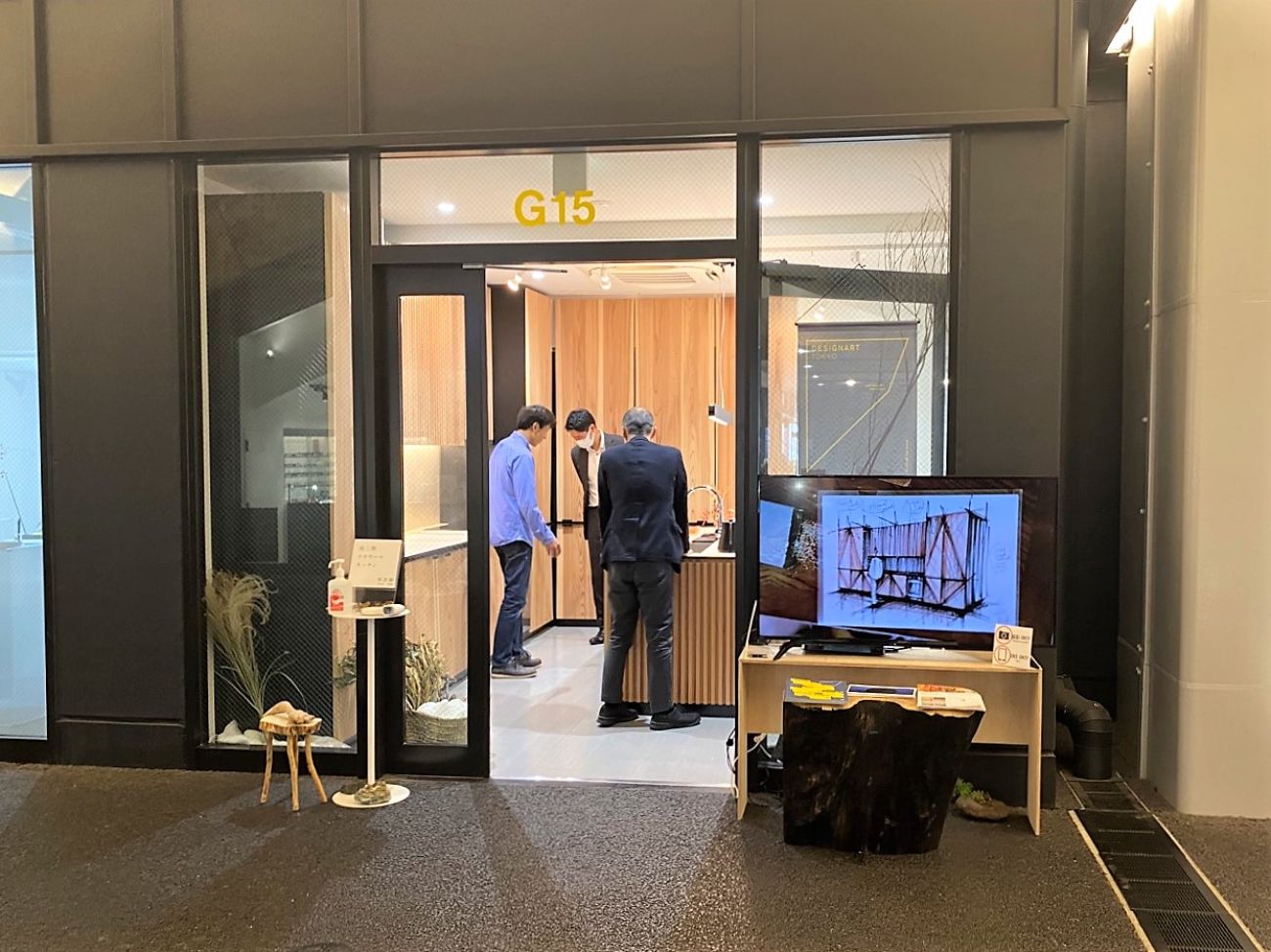 ガラス扉越しに木目調キッチンを見学する3人の姿と、外に設置された大型モニターが映る展示会場の写真