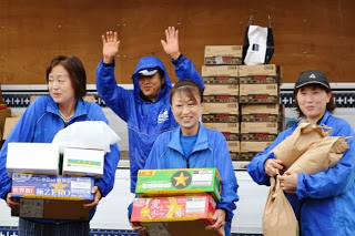 トラックの荷台の前で青い作業着を着た女性たちが笑顔で箱や紙袋に詰まった物資を抱えて喜んでいる様子を写した写真