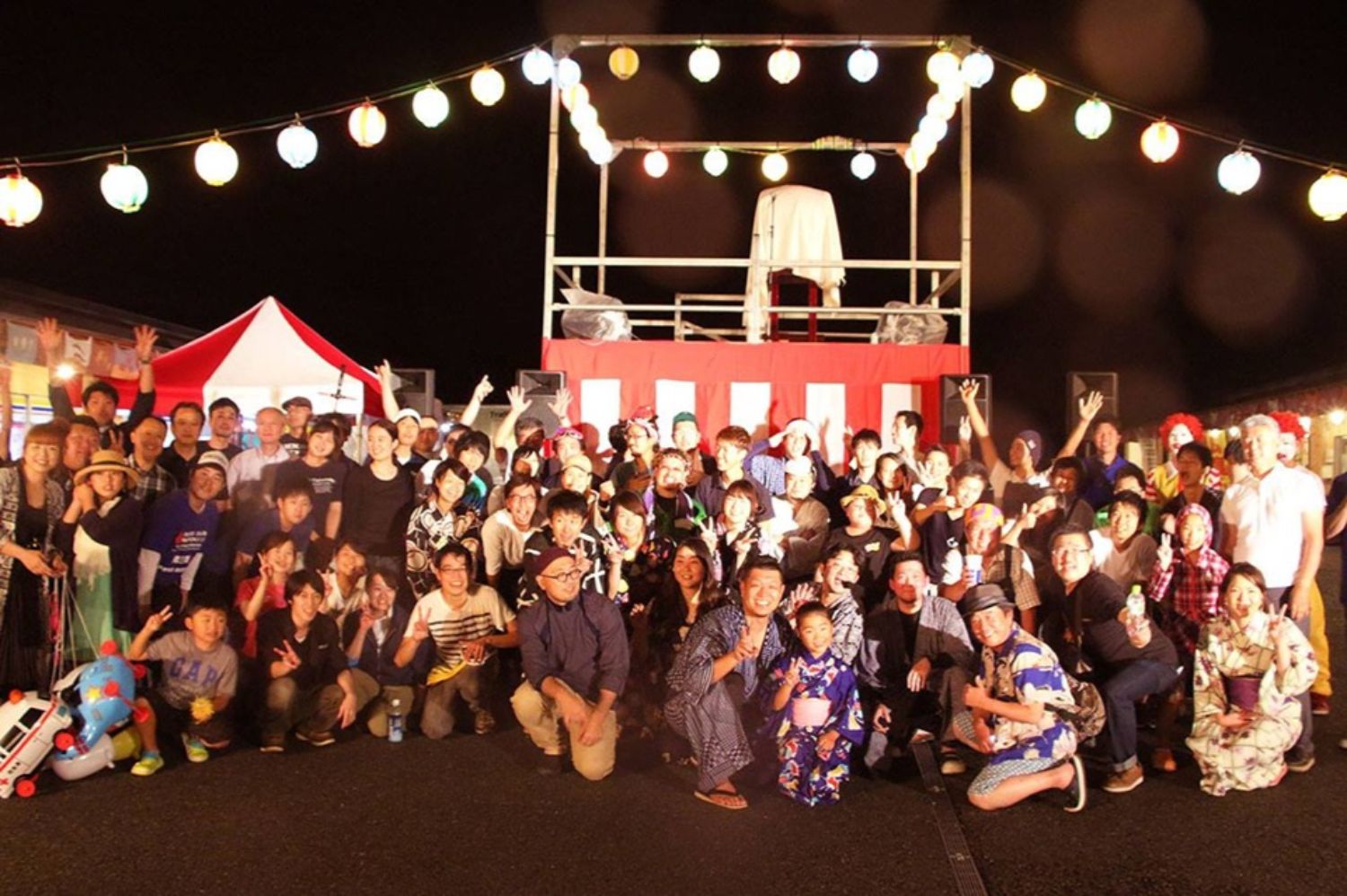 夜の盆踊り会場で、やぐらの前に浴衣姿や私服の人々が集まり、提灯の明かりに照らされながら笑顔で集合写真におさまっている様子を写した写真