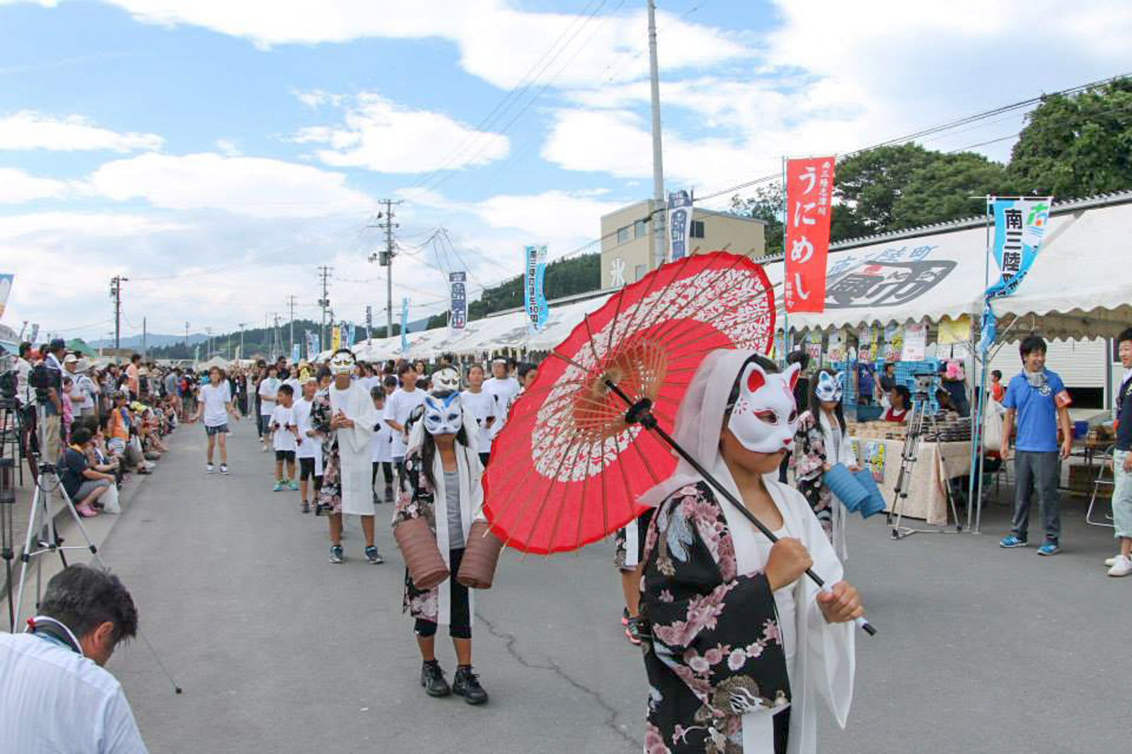 のぼりが立った白いテントが右側に立ち並び、左側に見物客が大勢集まっている道の中央を、赤い和傘を差し、着物柄の法被を着て狐の面をつけた人を先頭に、狐の面をつけた人々や子どもたちが行列になって歩いている様子の写真