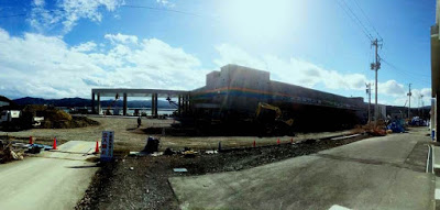 重機が作業している建設現場に、白い壁にオレンジ、緑、青の線がデザインされた新魚市場の施設が建っている写真