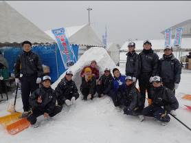 白いテントとイベント用の青いのぼり旗が立ち並ぶ会場で、雪で作ったかまくらの前に防寒着を着た子どもたちが集まり、笑顔でポーズをとっている写真