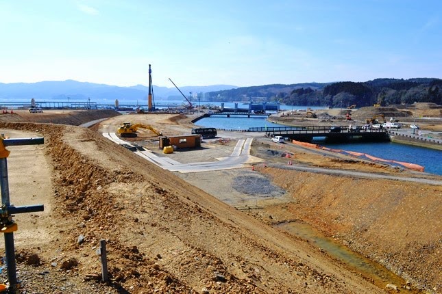 土を盛った広い造成地の中に水辺があり、奥には橋のような構造物と複数の重機が見え、港湾や堤防の工事が行われている写真