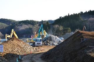 重機が土砂や石を積み上げ、中央にトラックが停まり、山林に囲まれた造成作業中の写真