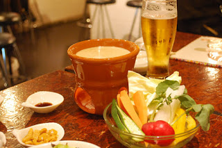 木のテーブルの上に、グラスに入ったビール、茶色のバーニャカウダポット、ラディッシュやオクラ、ニンジンなどの色とりどりの野菜が入った透明のボウルなどが並んでいる写真