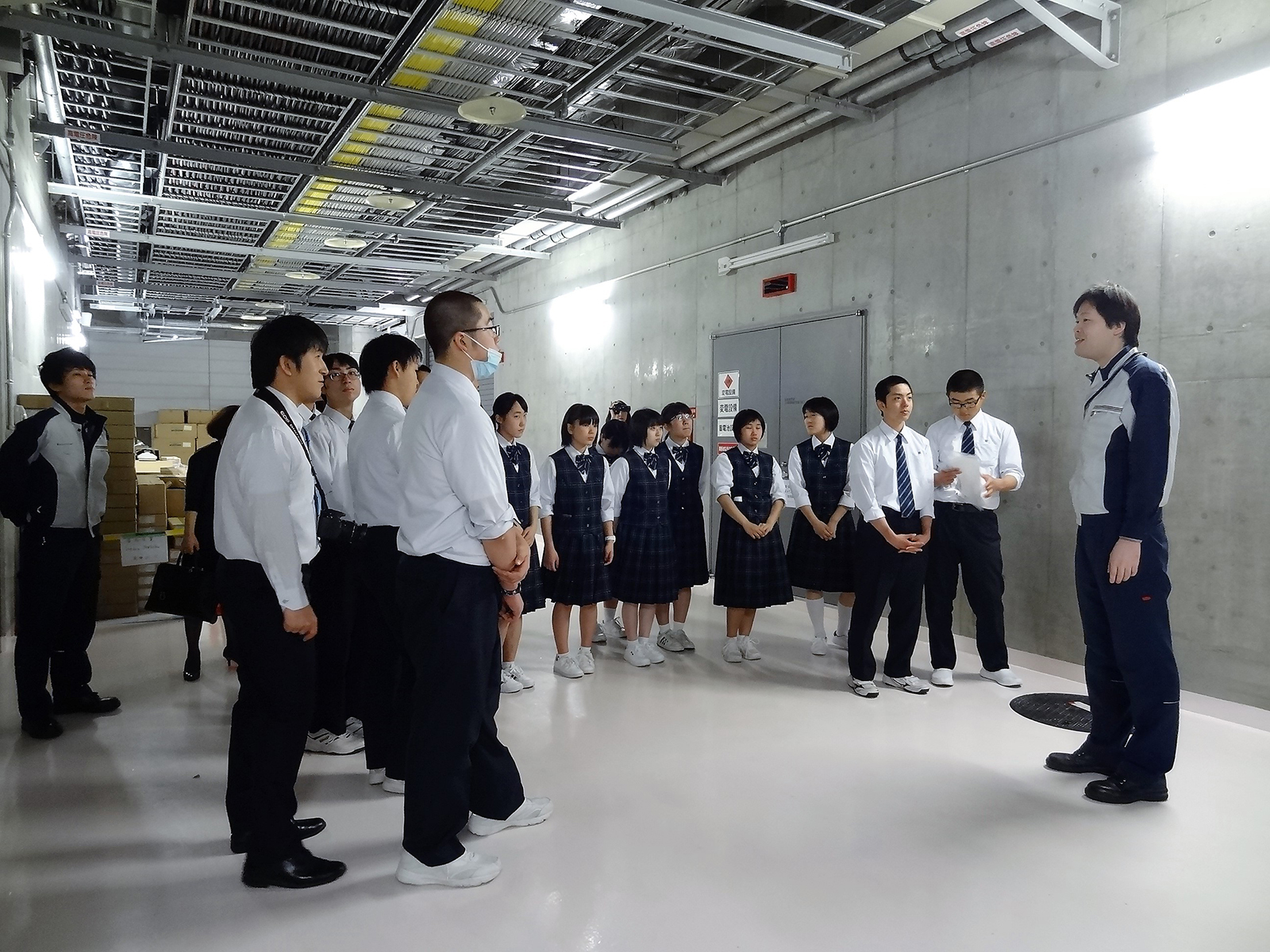 地下施設の通路で半円を描いて整列した高校生たちが担当者の男性の説明を聞きながら施設見学をしている写真