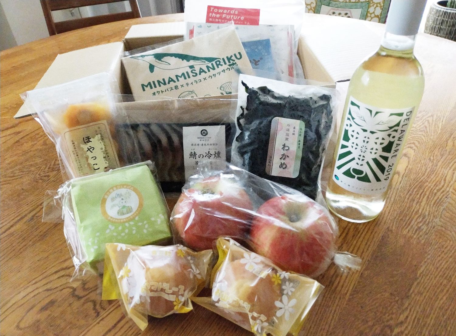 テーブルの上に、わかめや鯖の冷燻、ほや加工品、お菓子、リンゴ、ワインなど、南三陸をはじめとした地域の特産品がぎっしりと詰まった特産品セットが広げられている写真