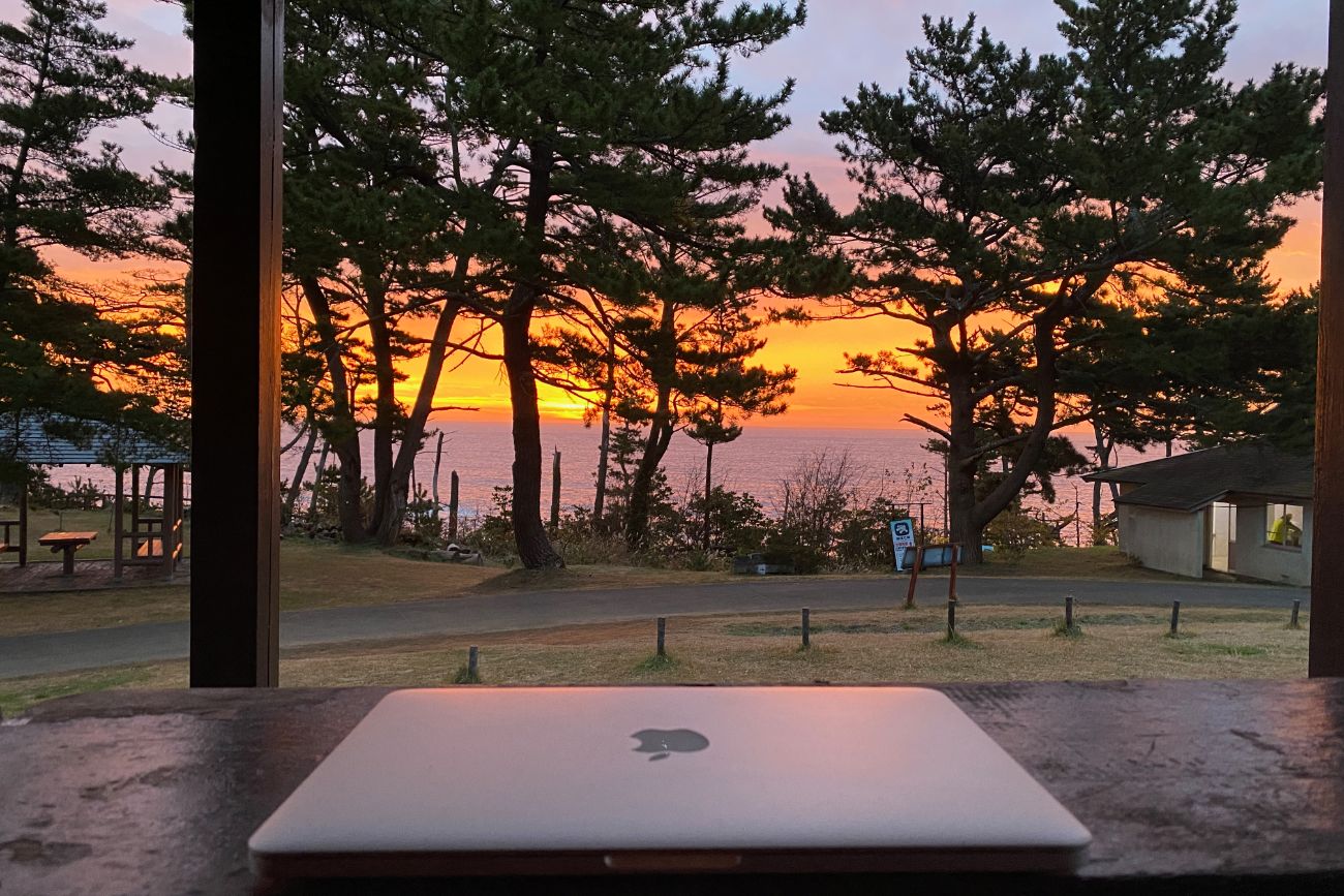 ベンチのある東屋や管理棟が見える松の木に囲まれたキャンプ場から見た夕焼けに染まった空と海の景色を、閉じられたノートパソコンが置かれている東屋から撮影した写真