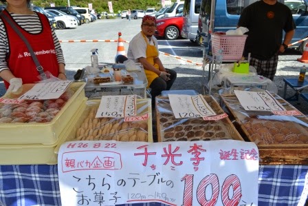 販売員が立ったり座ったりして笑顔を見せているお菓子の屋台で、値札の書かれた手書きの紙が並び、100円セールが行われているブースの写真