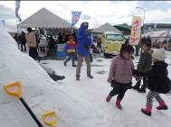 テントや屋台が並んだ会場の手前に作られた、雪の山に黄色い取っ手のスコップが立てかけられていて、そのそばを小さな子どもたちが楽しそうに歩いている様子の写真