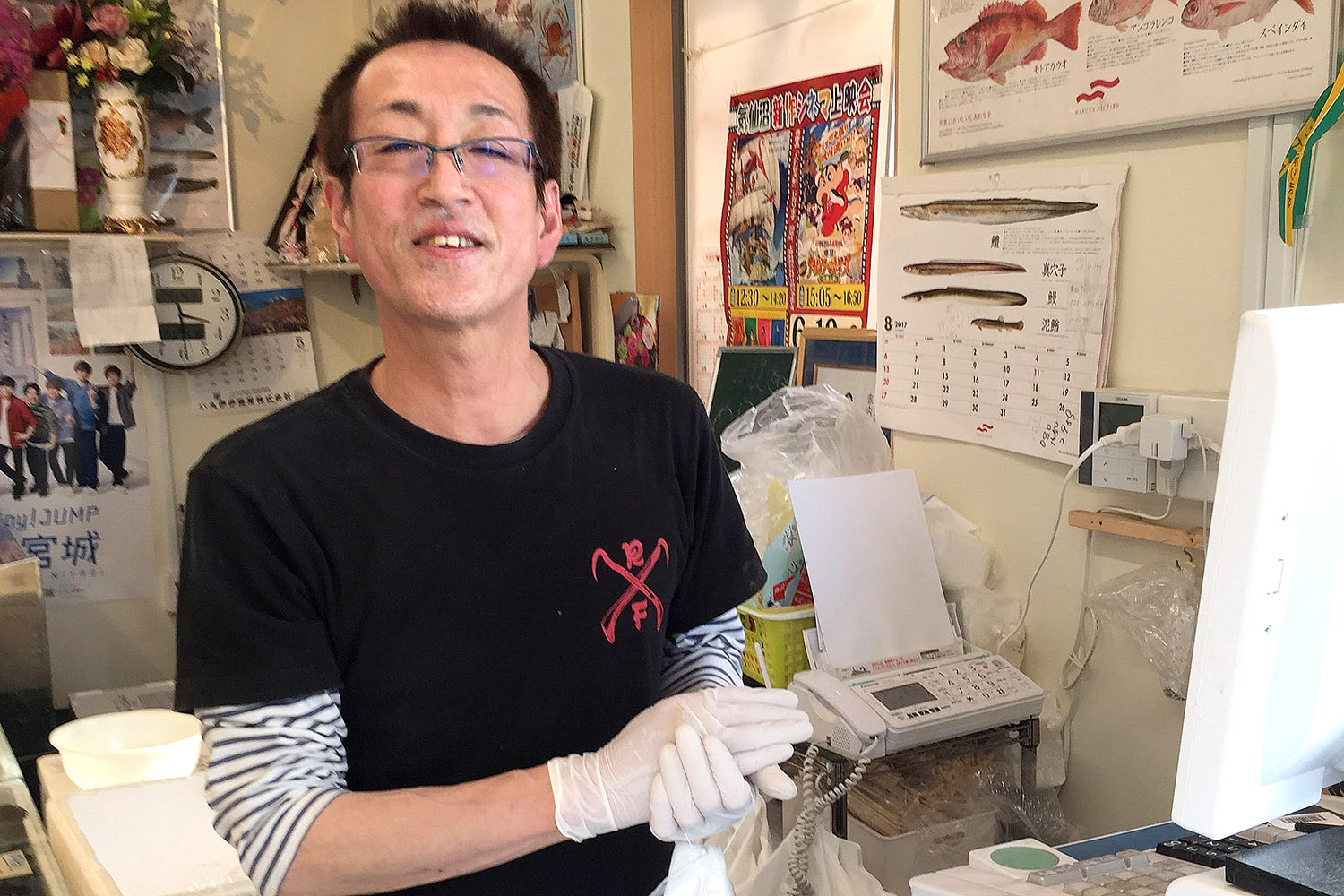 壁にポスターやカレンダーが貼られた店内に、白いゴム手袋をはめてカウンターの前に立つ、黒いTシャツを着たスタッフが真っすぐカメラを見ている写真