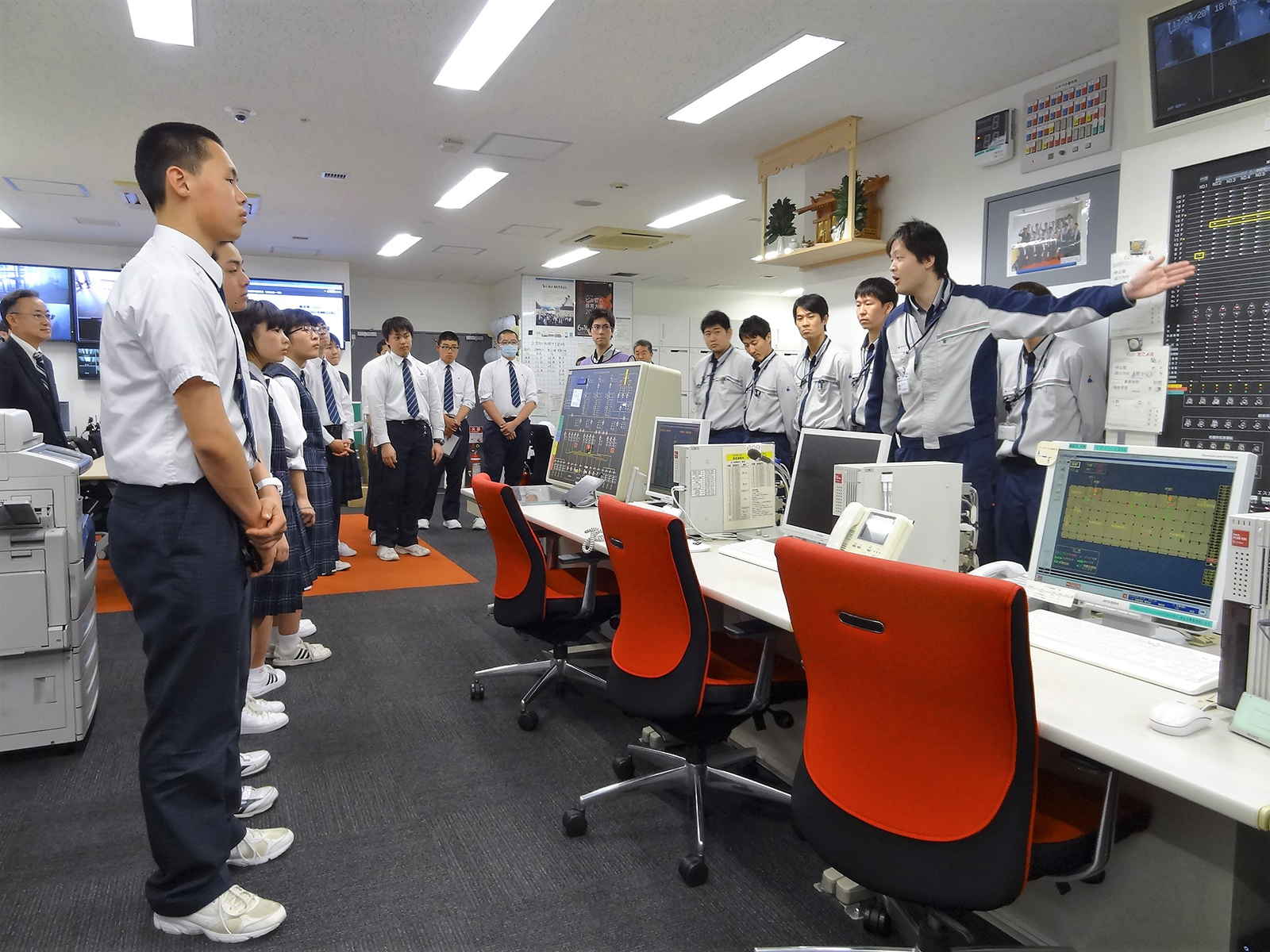 たくさんのモニターが壁やデスクに設置された防災センターで、制服姿の高校生たちが担当者の男性から説明を受けている写真