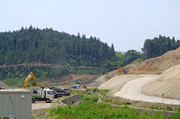 奥に緑の林が広がる茶色く削られた造成中の山の斜面を右側に、左側に並んだトラックと数台のショベルカーが作業を行っている工事現場の写真
