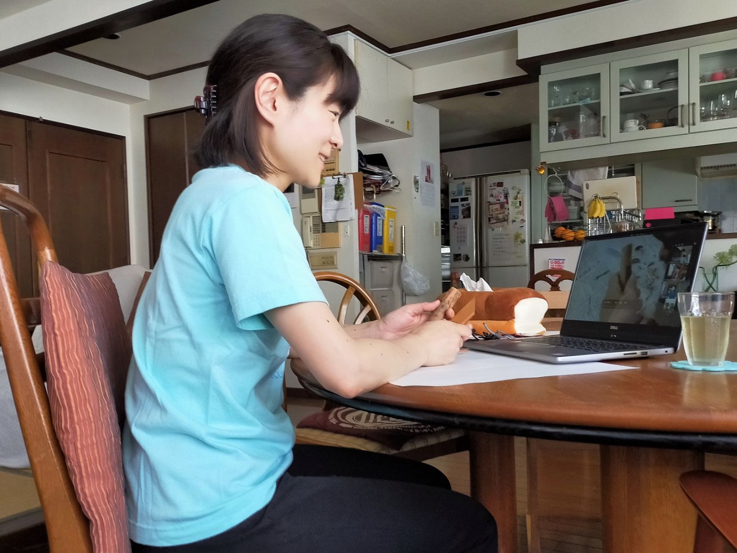 自宅のダイニングでパソコンのオンライン画面を見ながら、女性が木の枝と道具を手にカトラリー作りの作業をしている様子の写真