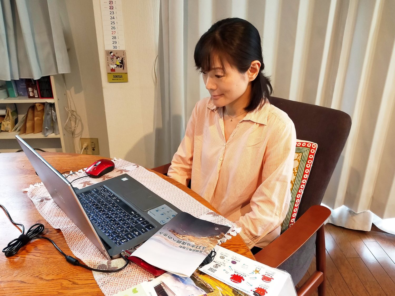 部屋の中で女性がノートパソコンの前に座り、資料やパンフレットを手元に置きながら、真剣な表情でオンラインツアーに参加している様子の写真