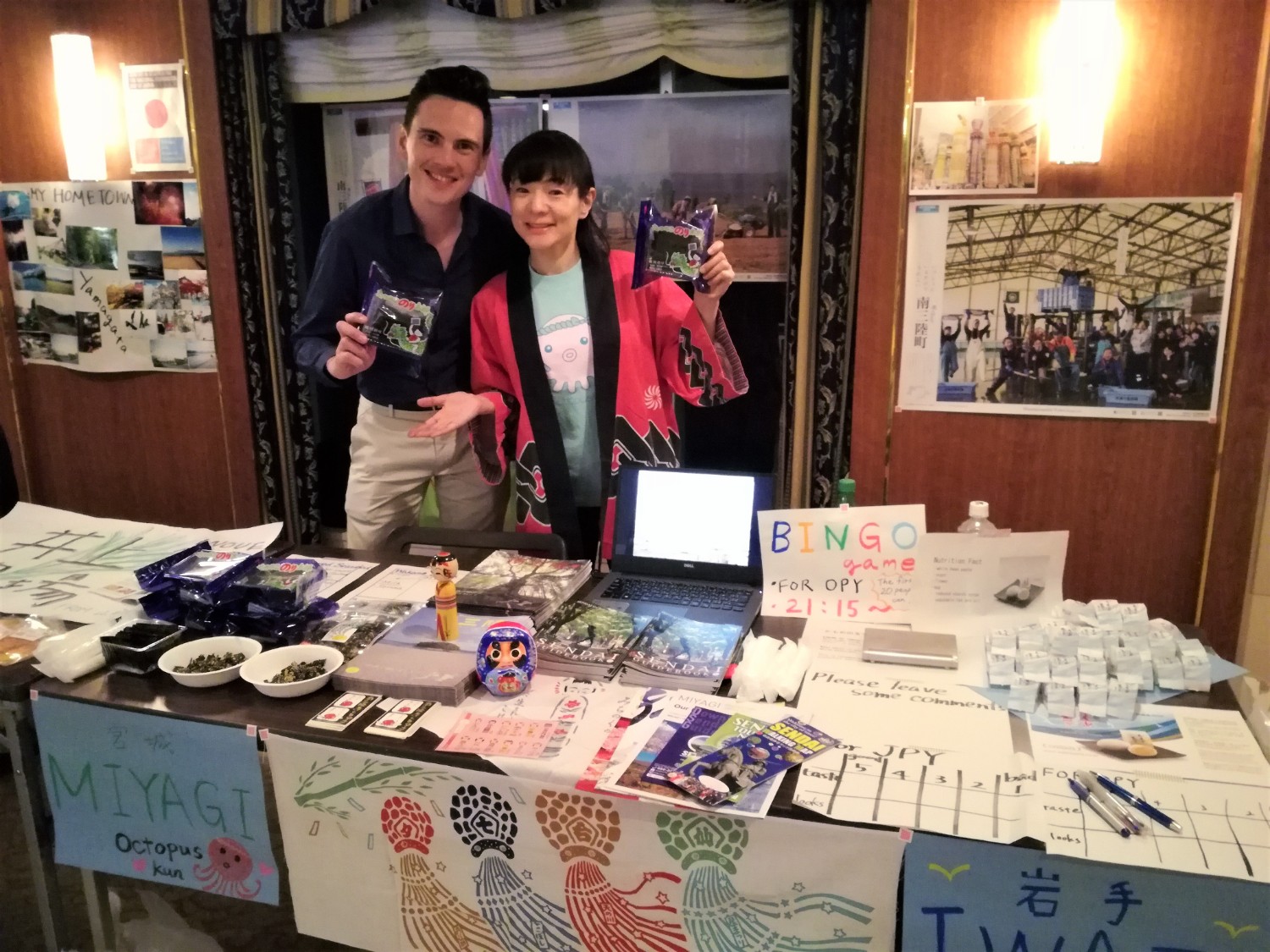 国際交流イベントで、日本の伝統的なはっぴを着た女性と外国人男性が、宮城県の特産品や観光情報を紹介するブースの前で笑顔で立ち、テーブルにはパンフレットや地元の食品などのが並んでいる様子の写真