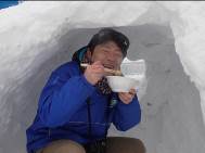 青い防寒着を着た男性が、雪で作ったかまくらの中に座り、手に持ったどんぶりから嬉しそうに食べ物を口に運んでいる様子の写真